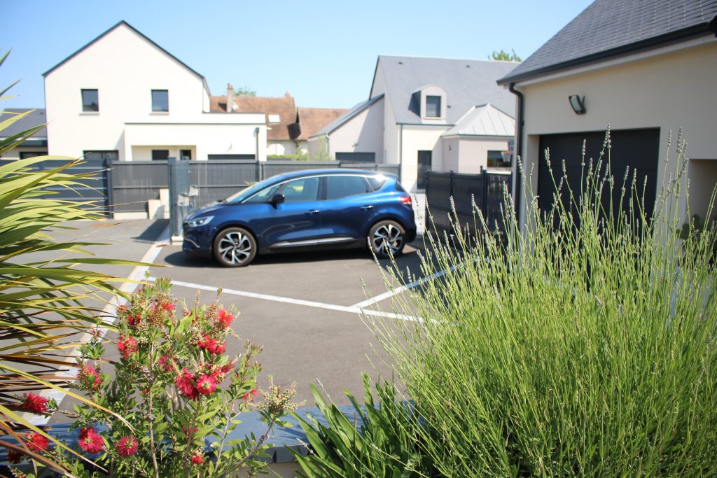 Accès carrossable en enrobé noir, paysagé par une jardinière pour cacher la zone de parking.