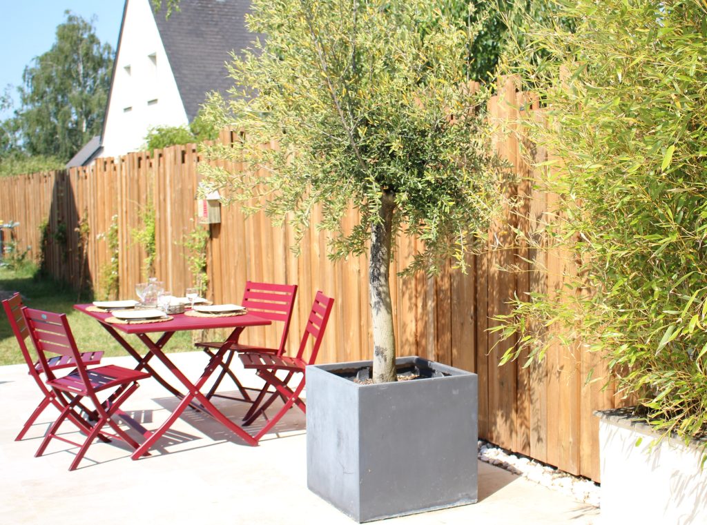 Terrasse décorée par du mobilier de jardin : une table en métal rouge et un bac carré gris avec un olivier.