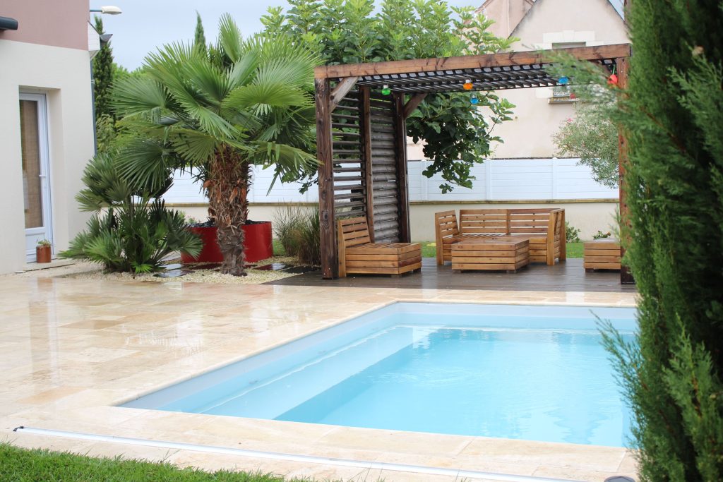 Terrasse en dalles de travertin et lames bois couverte d'une pergola et décorés par des massifs paysagers au bord d'une piscine.